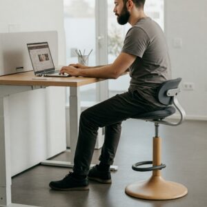 A person sitting comfortably on an ergonomic stool, working at a standing desk.
