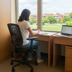A female student studying at a wooden desk in a dorm room, seated in a black mesh office chair with adjustable height and armrests.