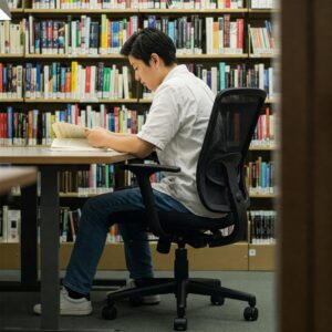 A student studying at a desk in a library, seated in a black mesh office chair with adjustable height and armrests, with bookshelves in the background.